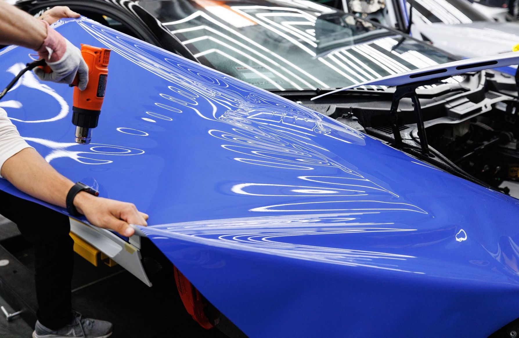 A technician uses a heat gun to carefully apply a blue vinyl wrap to a car in an auto workshop.